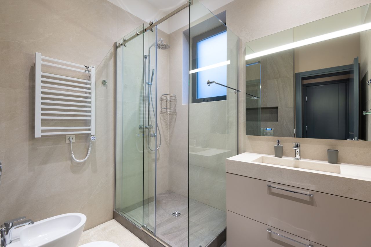 Shower cabin with glass walls near mirror hanging on wall above sink in modern minimalist styled bathroom